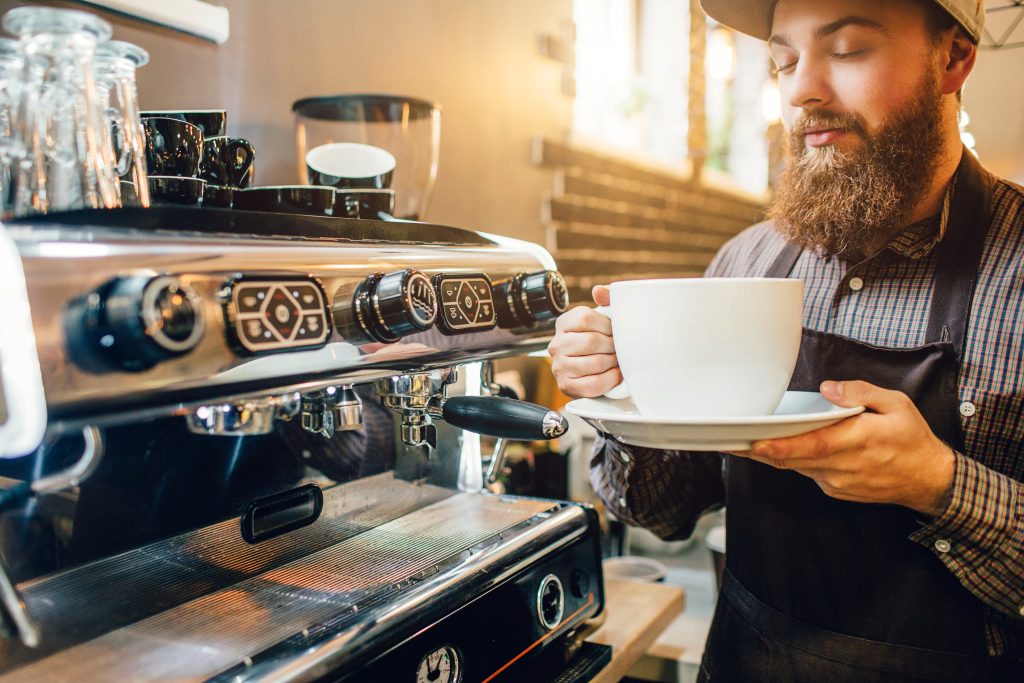 Calm and peaceful young man stand at coffee machine in kitchen and smells coffee from huge white cup. He looks at it with pleasure.